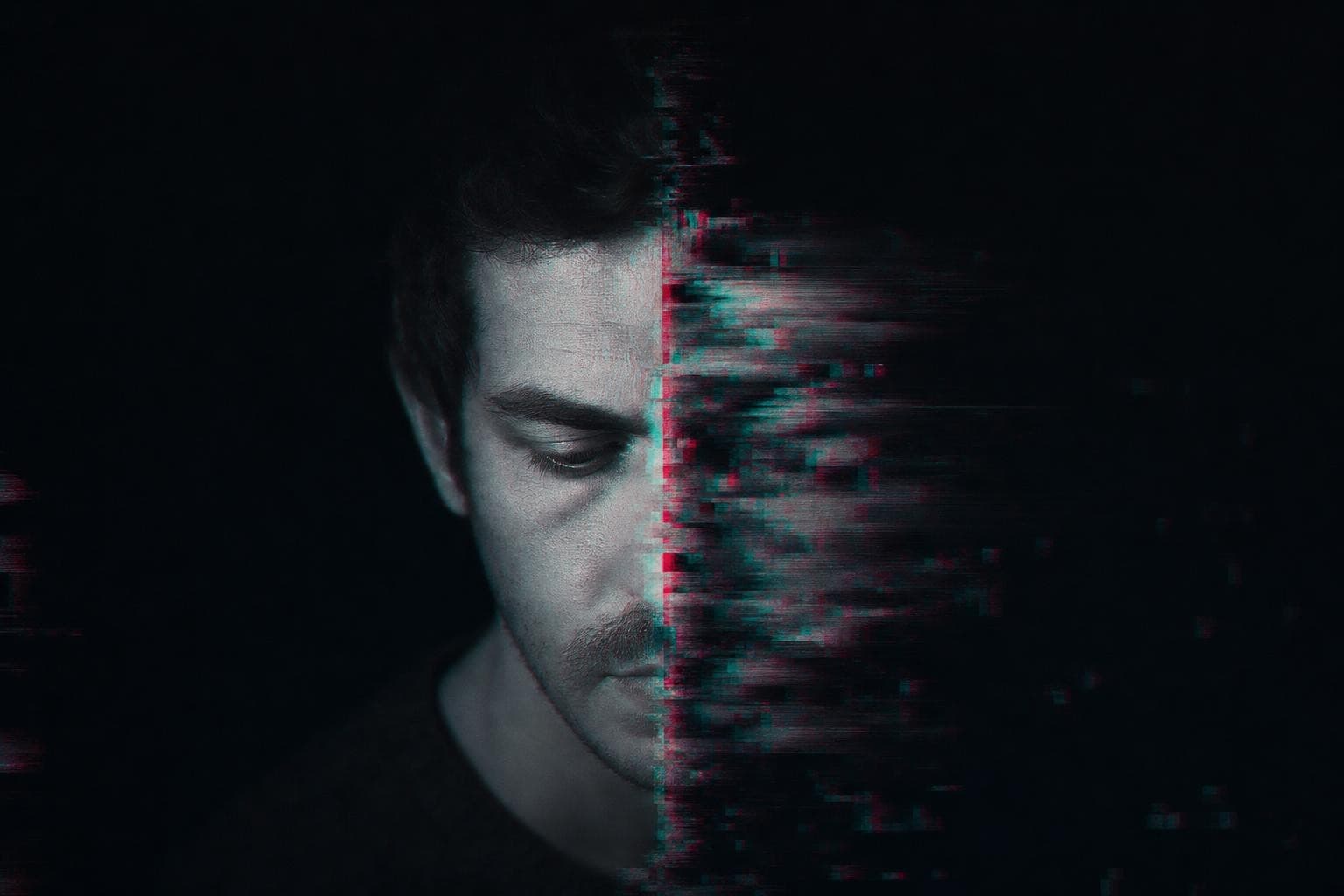 A close human portrait filling a near-black frame, split down the middle by a hard seam: the left half a crisp, cleanly captured face, the right half tearing apart into corrupted pixel blocks and a magenta-and-cyan RGB-split fringe, as if the same face cannot decide whether it is a real photograph or a synthetic one.
